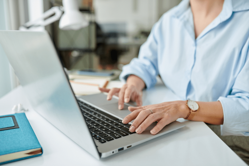 Persona seduta a una scrivania mentre utilizza un computer portatile, con mani sulla tastiera e ambiente di lavoro luminoso e ordinato.