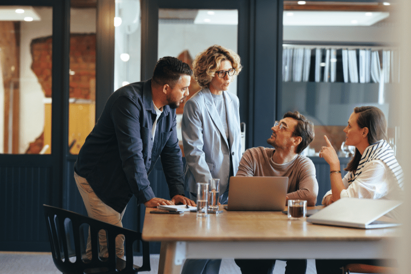 Quattro persone in piedi e sedute attorno a un tavolo discutono davanti a un computer portatile in un ufficio moderno.
