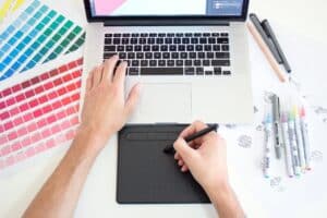 Designer visto dall'alto che lavora al colore con mazzette di campioni stampati, un laptop con software di grafica, una tavoletta grafica e schizzi a mano.