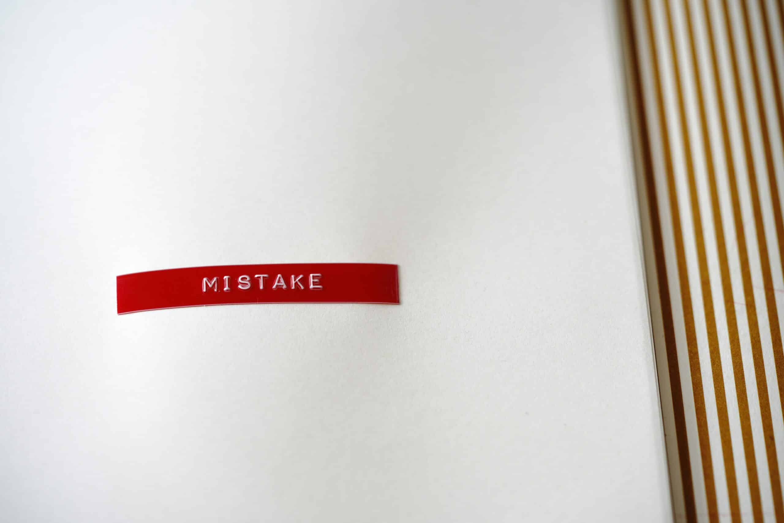 Etichetta rossa goffrata con la parola inglese mistake, cioè errore, su sfondo bianco accanto a strisce dorate verticali